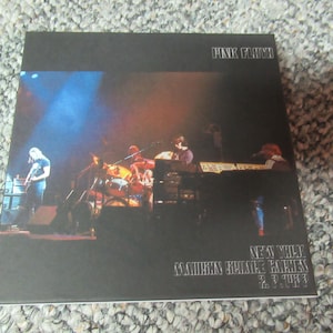 May include: A black and white album cover for Pink Floyd's "The Wall" album. The cover features a black and white photo of the band performing on stage. The album title is printed in white on the cover.