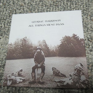 May include: A vintage vinyl record album cover featuring a sepia-toned photo of George Harrison sitting in a field with three other figures. The album title "George Harrison All Things Must Pass" is printed in white on a white background.