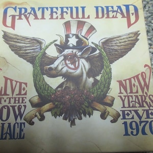 May include: Album cover for the Grateful Dead's "Live at the Cow Palace, New Year's Eve 1976." The artwork features a cow wearing an Uncle Sam hat and a monocle, with wings and a wreath. The album title and event details are in red and blue.