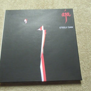 Puede incluir: Una portada de &aacute;lbum negra con la palabra "aja" en escritura roja y las palabras "Steely Dan" en blanco. Un dise&ntilde;o vertical de rayas rojas y blancas recorre el centro de la portada. El &aacute;lbum es cuadrado.
