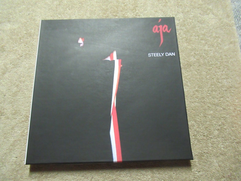 Puede incluir: Una portada de &aacute;lbum negra con la palabra "aja" en escritura roja y las palabras "Steely Dan" en blanco. Un dise&ntilde;o vertical de rayas rojas y blancas recorre el centro de la portada. El &aacute;lbum es cuadrado.