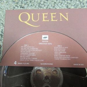 Puede incluir: Una cinta de carrete Queen Greatest Hits cuadrafónica vintage. La portada del álbum es de color marrón rojizo apagado con el nombre de la banda en letras doradas. La lista de canciones incluye éxitos como "We Will Rock You". Hecho en EE. UU.