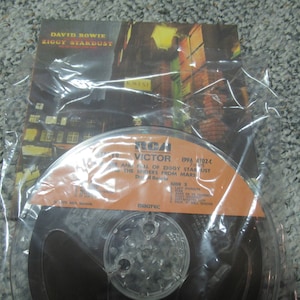 May include: A vintage audio reel tape of David Bowie's album, Ziggy Stardust, in a plastic sleeve. The album cover art features a nighttime city scene. The tape reel has an orange label with the RCA Victor logo.