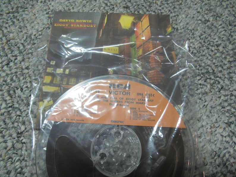 May include: A vintage audio reel tape of David Bowie's album, Ziggy Stardust, in a plastic sleeve. The album cover art features a nighttime city scene. The tape reel has an orange label with the RCA Victor logo.