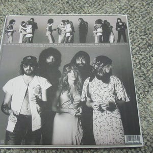 Puede incluir: Portada de &aacute;lbum de fotos en blanco y negro con una banda de siete personas. La portada del &aacute;lbum tiene el nombre de la banda "Fleetwood Mac" impreso en letras blancas en la parte superior. El t&iacute;tulo del &aacute;lbum "Rumours" est&aacute; impreso en letras blancas debajo del nombre de la banda. La portada del &aacute;lbum presenta una foto de los miembros de la banda de pie en fila. Los miembros de la banda visten ropa informal. La portada del &aacute;lbum es un ejemplo cl&aacute;sico del arte de las portadas de &aacute;lbumes de la d&eacute;cada de 1970.