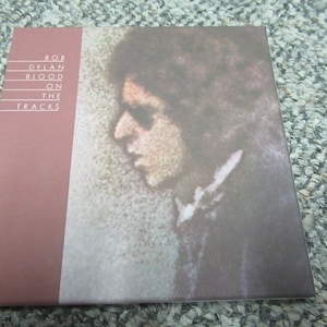 May include: A red album cover with the text "BOB DYLAN BLOOD ON THE TRACKS" in white. The cover features a close-up of a person's face, with a soft, watercolor-like style.