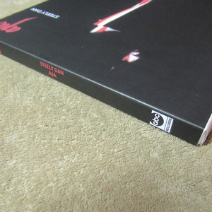 Puede incluir: Primer plano de la portada del &aacute;lbum "Aja" de Steely Dan. La portada es negra con texto rojo y una imagen parcial. El logotipo de ABC Records es visible en la esquina inferior derecha. El &aacute;lbum descansa sobre una superficie beige.