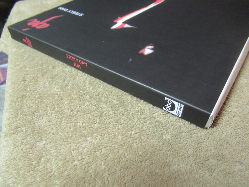 Puede incluir: Primer plano de la portada del &aacute;lbum "Aja" de Steely Dan. La portada es negra con texto rojo y una imagen parcial. El logotipo de ABC Records es visible en la esquina inferior derecha. El &aacute;lbum descansa sobre una superficie beige.