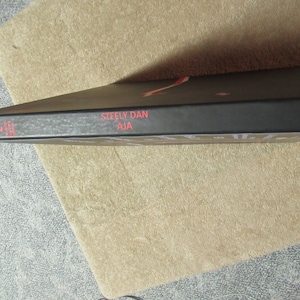 Puede incluir: Una portada de &aacute;lbum negra con el texto "Steely Dan Aja" en rojo. El logotipo de ABC Records es visible en el lado derecho. El &aacute;lbum est&aacute; sobre una superficie beige.