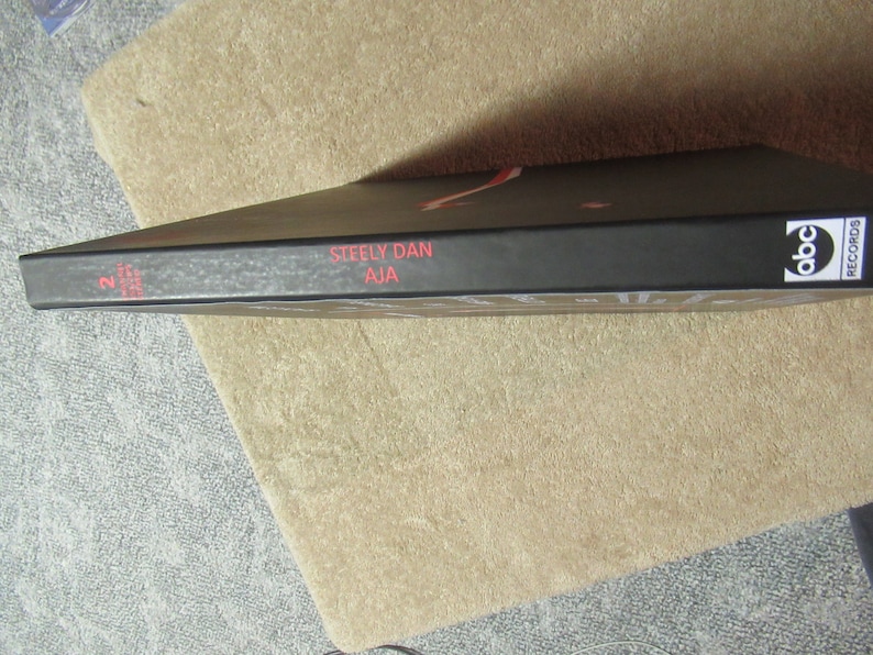 Puede incluir: Una portada de &aacute;lbum negra con el texto "Steely Dan Aja" en rojo. El logotipo de ABC Records es visible en el lado derecho. El &aacute;lbum est&aacute; sobre una superficie beige.