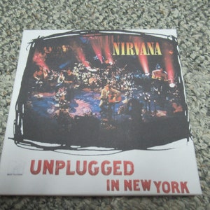 Könnte beinhalten: Ein weißes CD-Gehäuse mit einem Schwarzweißfoto der Band Nirvana, die live auf der Bühne spielt. Der Text "NIRVANA" ist oben auf dem Gehäuse in Gelb. Der Text "UNPLUGGED IN NEW YORK" ist unten auf dem Gehäuse in Rot.