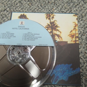 May include: A vintage audio reel-to-reel tape and album cover for the Eagles' "Hotel California." The tape is in a clear plastic case, and the album cover features a sunset scene with palm trees. The text on the tape reads "Eagles Hotel California."