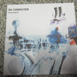 Könnte beinhalten: Ein CD-Cover für das Album "OK Computer" von Radiohead. Das Cover zeigt ein blaues und weißes abstraktes Design mit einem Baum im Hintergrund und dem Albumtitel und dem Künstlernamen in weißer Schrift.