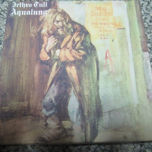 May include: A vintage vinyl record album cover featuring a painting of a man in a brown coat with long hair and a beard. The album cover is for the Jethro Tull album "Aqulung" and features the text "Aqualung" in large, bold letters at the top of the cover. The album cover also features the text "The  Christmas Song" in smaller letters at the top right corner of the cover.