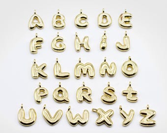 May include: A collection of gold-colored alphabet charms, each with a loop for attaching to a necklace or bracelet. The letters are in a rounded, bubble-like font, and are arranged in rows against a white background.