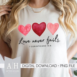 May include: A white t-shirt with three watercolor hearts in shades of pink and red. The text "Love never fails." is printed below the hearts, with the scripture reference "1 Corinthians 13:8" underneath.