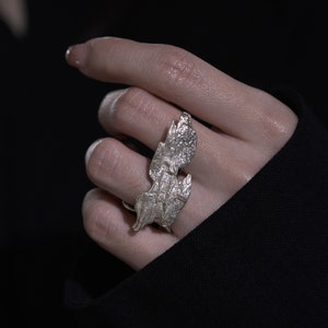 May include: A silver ring with a textured, organic design resembling two overlapping leaves. The ring is worn on a finger.