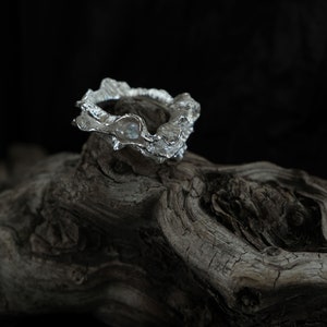 Puede incluir: Un anillo de plata con un diseño texturizado y orgánico. El anillo está apoyado en un trozo de madera flotante desgastada.