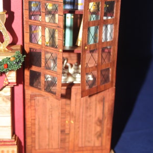 May include: A wooden cabinet with glass doors and shelves. The cabinet is painted a dark brown color and has a distressed finish. The glass doors have a diamond pattern and are open, revealing shelves filled with books and other items. The cabinet is sitting on a wooden floor.