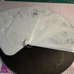 Könnte beinhalten: Ein Satz weißer Kunststoffschablonen mit verschiedenen Designs, darunter Wirbel und andere Formen, die durch einen Metallring zusammengehalten werden. Die Schablonen sind auf einer dunklen Oberfläche aufgefächert, mit einer Schneidematte im Hintergrund.