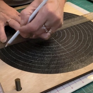 Op de afbeelding: Een houten knutselbord met een zwart cirkelvormig ontwerp en concentrische cirkels. Een liniaal en potlood worden gebruikt om lijnen op het bord te tekenen. Het bord heeft een lichte houtkleur en wordt gebruikt voor knutselen.