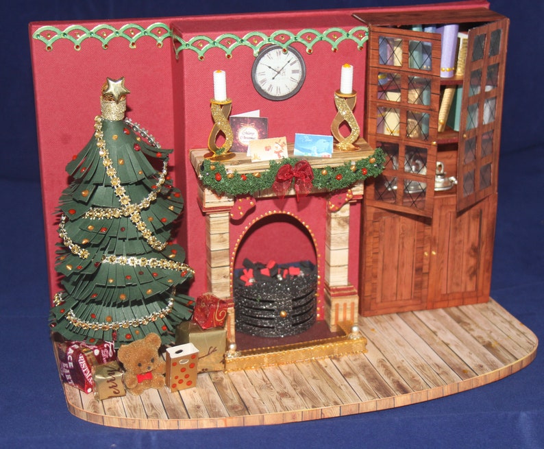 May include: A miniature papercraft Christmas scene with a fireplace, a decorated Christmas tree, a bookcase, and presents under the tree.