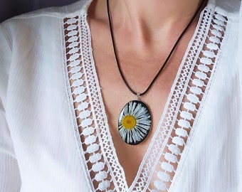 May include: Oval pendant necklace with a pressed daisy, yellow centre and white petals, set in a silver frame. The pendant hangs from a black cord. A piece of floral jewellery.