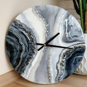 May include: A round, white, gray, and black resin clock with a black clock hand and a white background. The clock face has a swirling, abstract design with silver glitter accents.