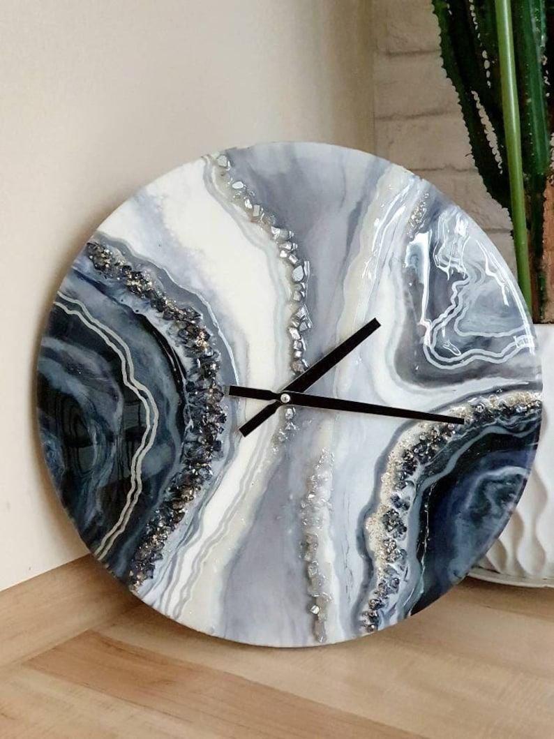May include: A round, white, gray, and black resin clock with a black clock hand and a white background. The clock face has a swirling, abstract design with silver glitter accents.