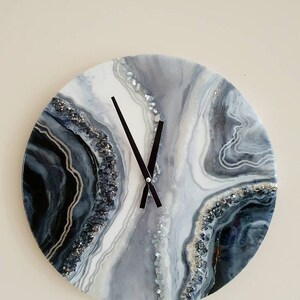 May include: A round, white, gray, and black resin clock with a black clock face and black hands. The clock face has a swirling, abstract design with silver glitter embedded in the resin.