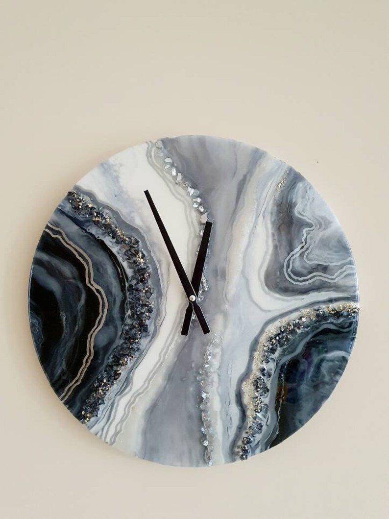 May include: A round, white, gray, and black resin clock with a black clock face and black hands. The clock face has a swirling, abstract design with silver glitter embedded in the resin.