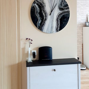 May include: A white dresser with two drawers and black handles. A black and white abstract clock with silver glitter is hanging on the wall above the dresser. A black speaker is on top of the dresser.