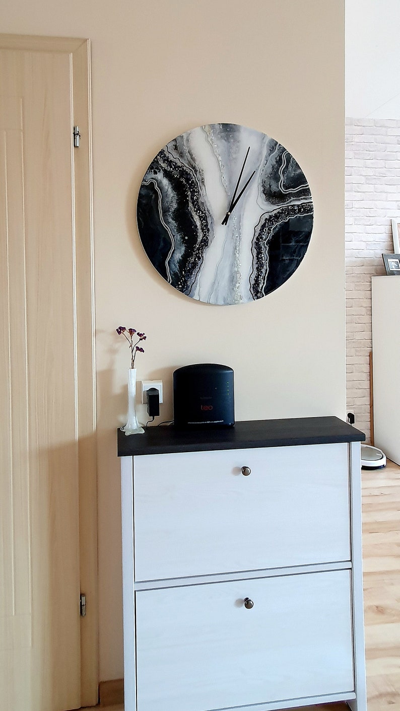 May include: A white dresser with two drawers and black handles. A black and white abstract clock with silver glitter is hanging on the wall above the dresser. A black speaker is on top of the dresser.