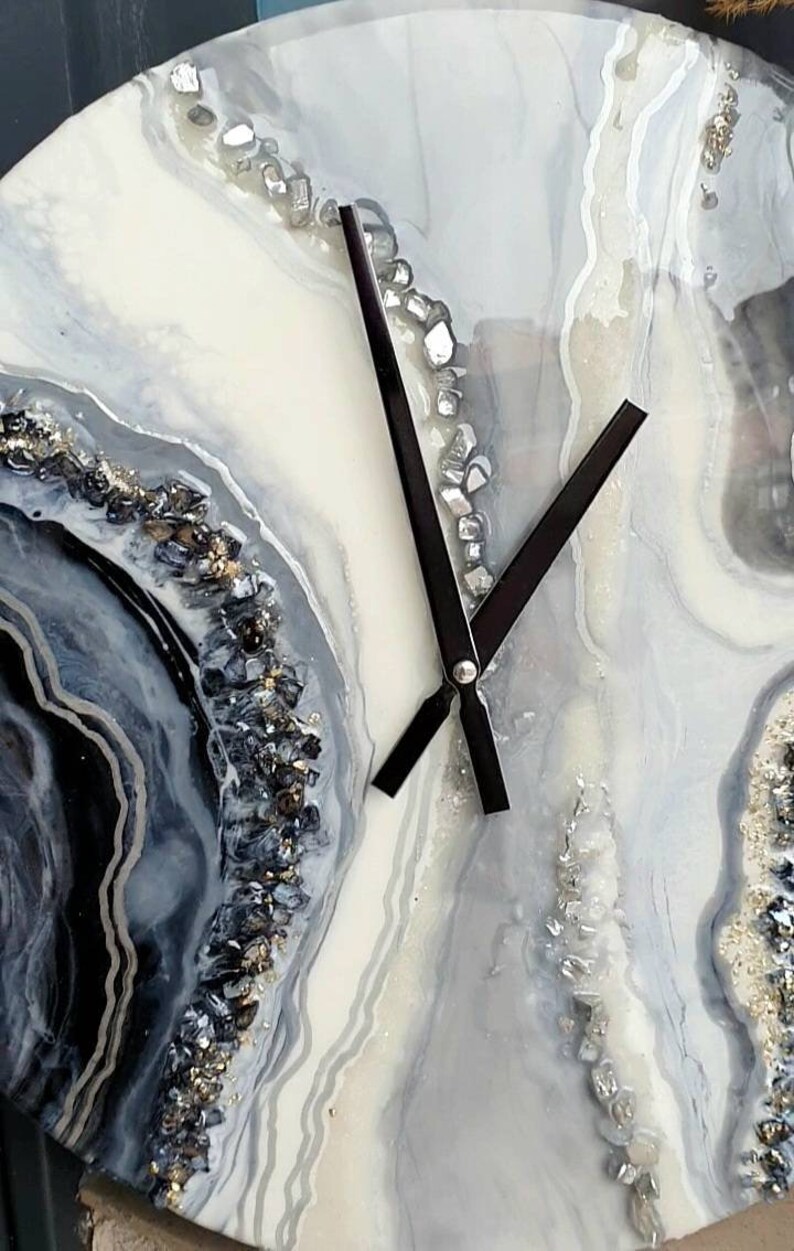 May include: A white and gray resin clock with black hands and silver glitter accents. The clock face features a swirling agate design.