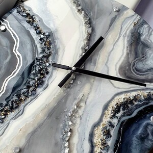 May include: A round, white and gray resin clock with black hands. The clock face has a swirling, abstract design with silver and gold glitter embedded in the resin.