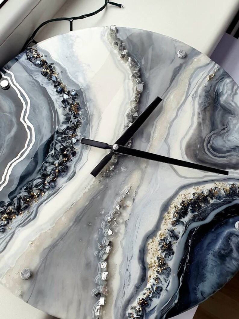 May include: A round, white and gray resin clock with black hands. The clock face has a swirling, abstract design with silver and gold glitter embedded in the resin.