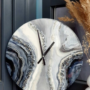 May include: A round, white, gray, and black resin clock with a black clock face and silver glitter accents. The clock has a unique, abstract design that resembles agate.