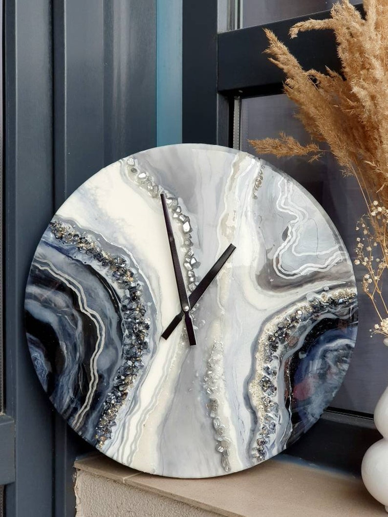 May include: A round, white, gray, and black resin clock with a black clock face and silver glitter accents. The clock has a unique, abstract design that resembles agate.