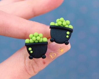 May include: A pair of black cauldron stud earrings with green bubbling potion. The earrings are held in a hand against a blue background. Each cauldron has three small yellow dots and three black feet.