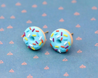 May include: A pair of donut-shaped stud earrings. Each earring features a light blue frosting with colorful sprinkles. The earrings are set against a light blue background with a repeating pattern of small pink shapes.