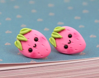 May include: A pair of pink strawberry earrings with green leaves and a smiling face. The earrings are on a blue book with a white cover.