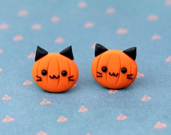 May include: A pair of orange pumpkin-shaped earrings with black cat ears and faces. The earrings are on a blue background with a white polka dot pattern.