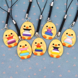 Może przedstawiać: Siedem żółtych breloków w kształcie kurczaka z różnymi flagami dumy. Każdy brelok ma czarny sznurek ze srebrnym zapięciem.