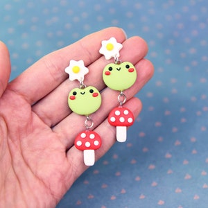 May include: A pair of whimsical dangle earrings featuring a light green frog with rosy cheeks, a red mushroom with white spots, and a white flower with a yellow center. The earrings are connected by silver-colored metal. The earrings are held in a hand.