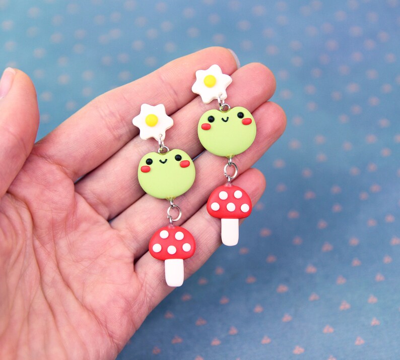 May include: A pair of whimsical dangle earrings featuring a light green frog with rosy cheeks, a red mushroom with white spots, and a white flower with a yellow center. The earrings are connected by silver-colored metal. The earrings are held in a hand.