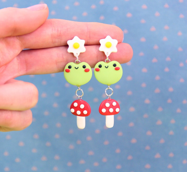 May include: A pair of whimsical dangle earrings featuring a daisy, a smiling green frog, and a red mushroom with white spots. The earrings are held up by a hand against a blue background with white dots.