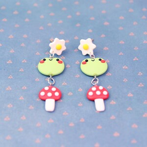 May include: A pair of whimsical earrings featuring a light green frog with rosy cheeks, a red mushroom with white spots, and a white flower with a yellow center. The earrings are connected with silver-colored metal. The background is a light blue surface with a pink pattern.