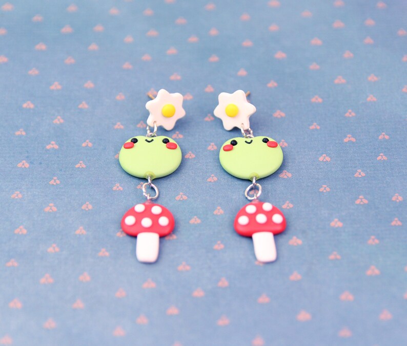 May include: A pair of whimsical earrings featuring a light green frog with rosy cheeks, a red mushroom with white spots, and a white flower with a yellow center. The earrings are connected with silver-colored metal. The background is a light blue surface with a pink pattern.