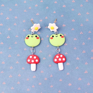 May include: A pair of whimsical earrings featuring a green frog, a red mushroom with white spots, and a white flower with a yellow center. The earrings are connected with silver-colored metal. The background is a light blue fabric with a pink floral pattern.