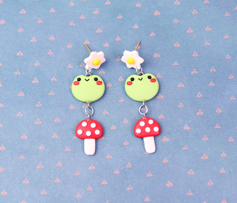May include: A pair of whimsical earrings featuring a green frog, a red mushroom with white spots, and a white flower with a yellow center. The earrings are connected with silver-colored metal. The background is a light blue fabric with a pink floral pattern.